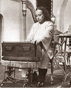 Black and white photo of Paramahansa Yogananda seated playing a harmonium on a small round table.