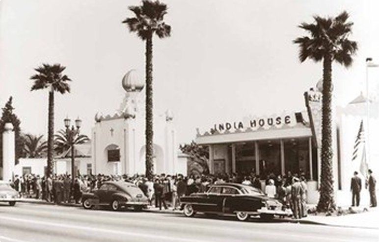 Vintage photo of a crowd outside India House with classic cars and tall palm trees along the street.
