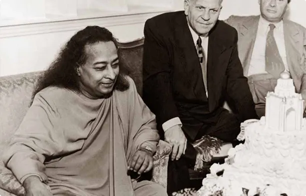 Paramahansa Yogananda sitting with two men in suits near an elaborate cake.
