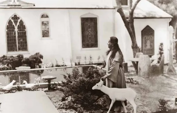 Paramahansa Yogananda standing outdoors near a white deer and a church with stained-glass windows.