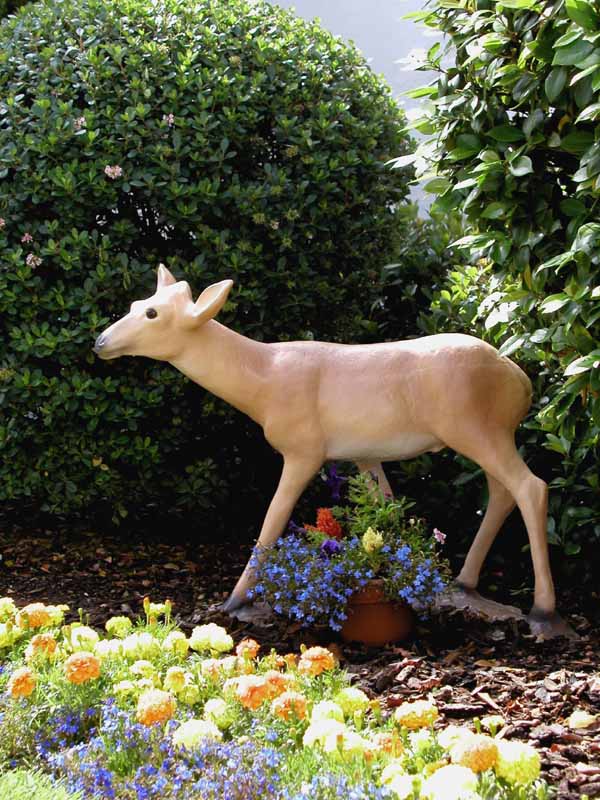 Deer statue standing in a garden surrounded by flowers and green bushes.