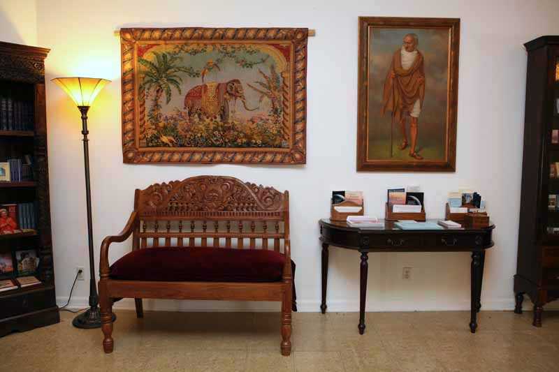 Room interior with carved wooden bench, tapestry with elephant, lamp, portrait, and table with brochures.
