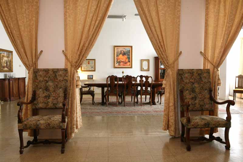 Room with patterned armchairs, gold curtains, and wooden chairs around a table under a framed portrait.
