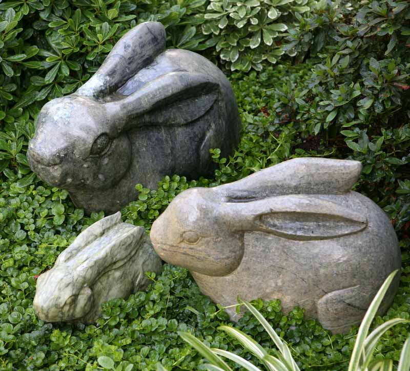 Three stone rabbit sculptures nestled among green leafy plants in a garden setting.