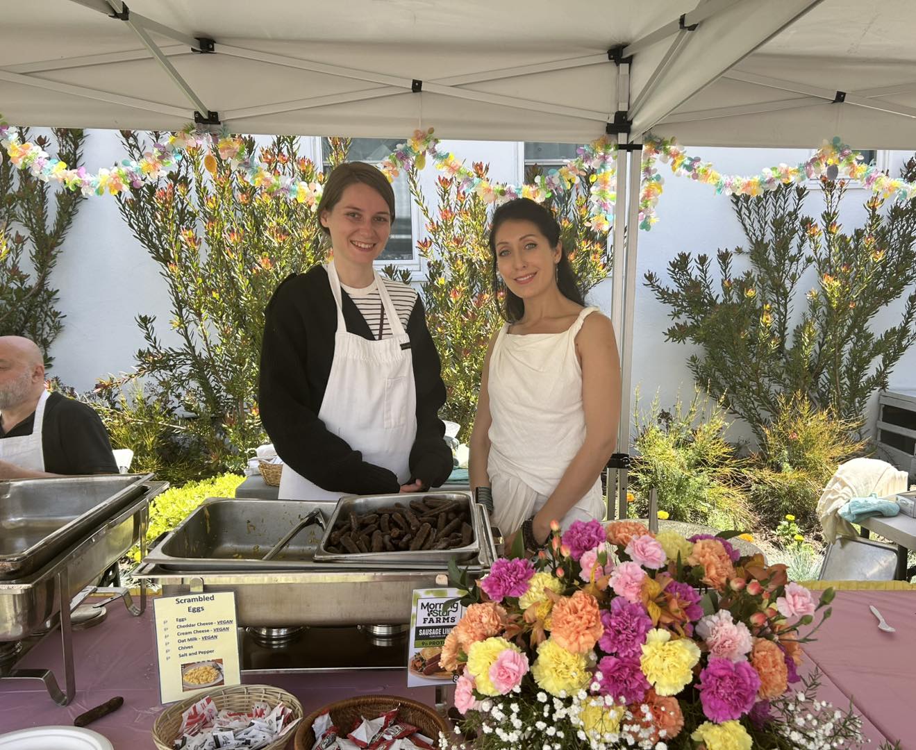 Two women smiling behind a buffet table with breakfast food trays and colorful flowers under a white canopy.