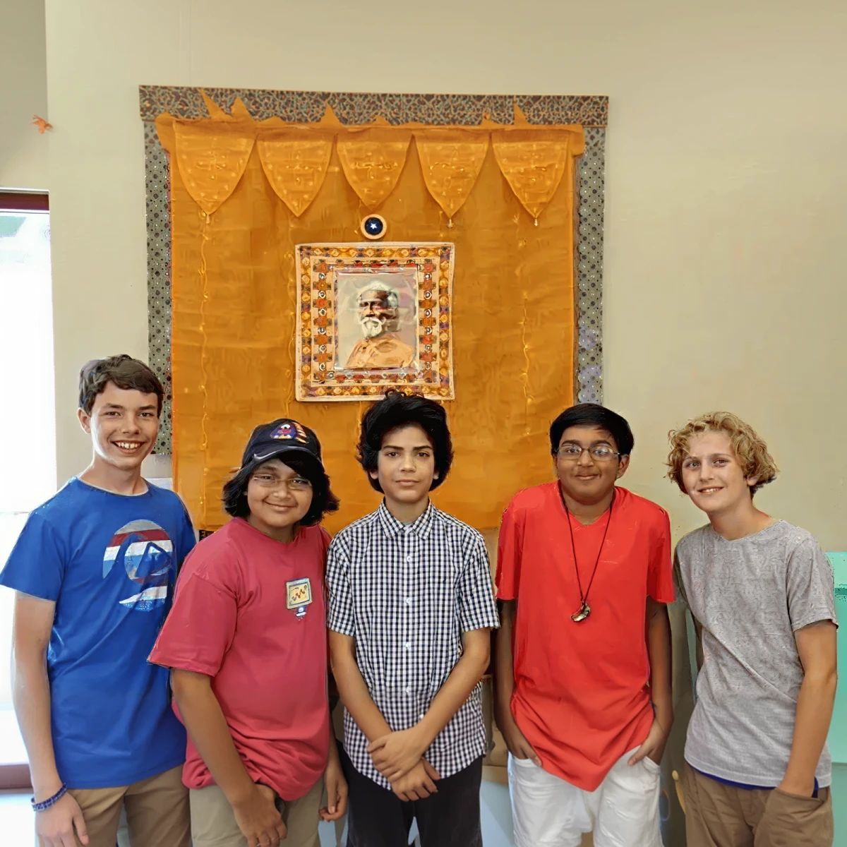 Group of five diverse boys standing indoors in front of a yellow and orange decorative wall hanging with a framed portrait.