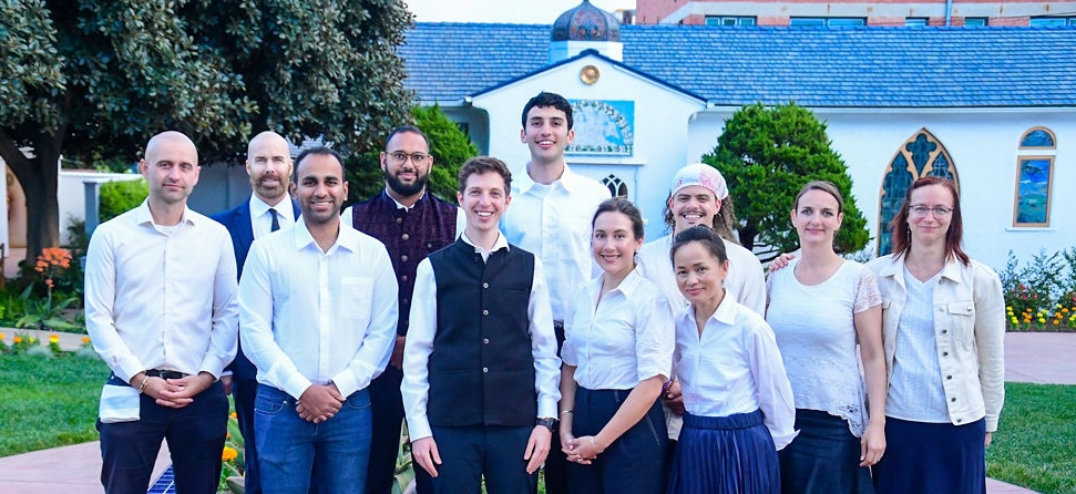 Group of eleven adults standing outdoors in front of a white building with blue roof and trees.