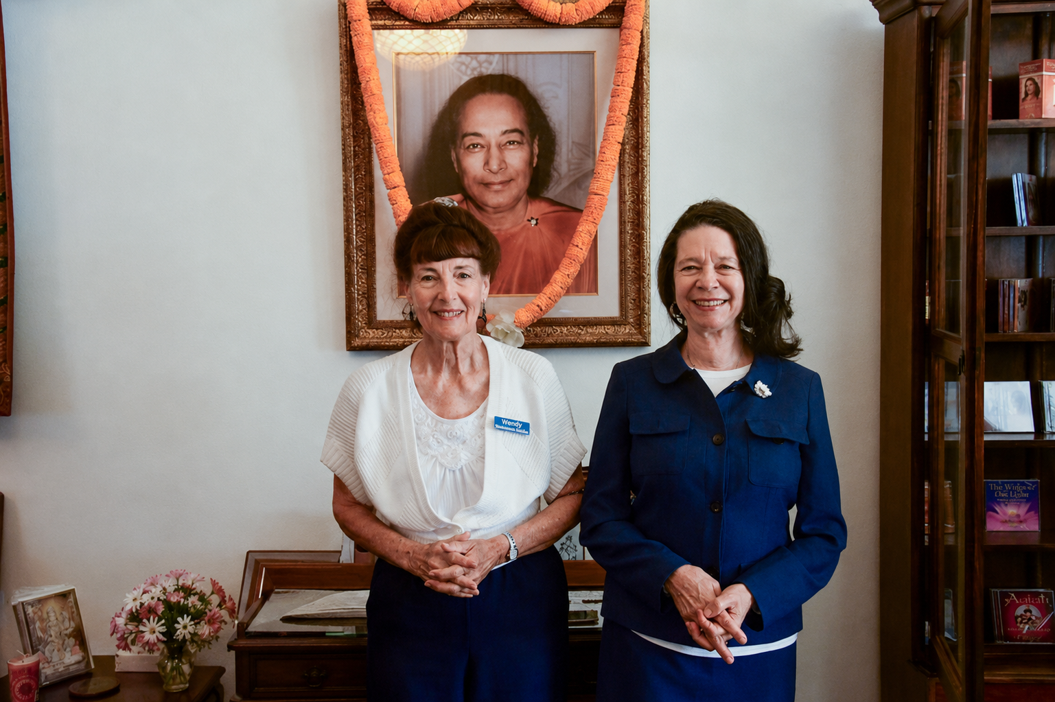 Three smiling women standing indoors in front of a framed portrait of Paramahansa Yogananda decorated with a garland.