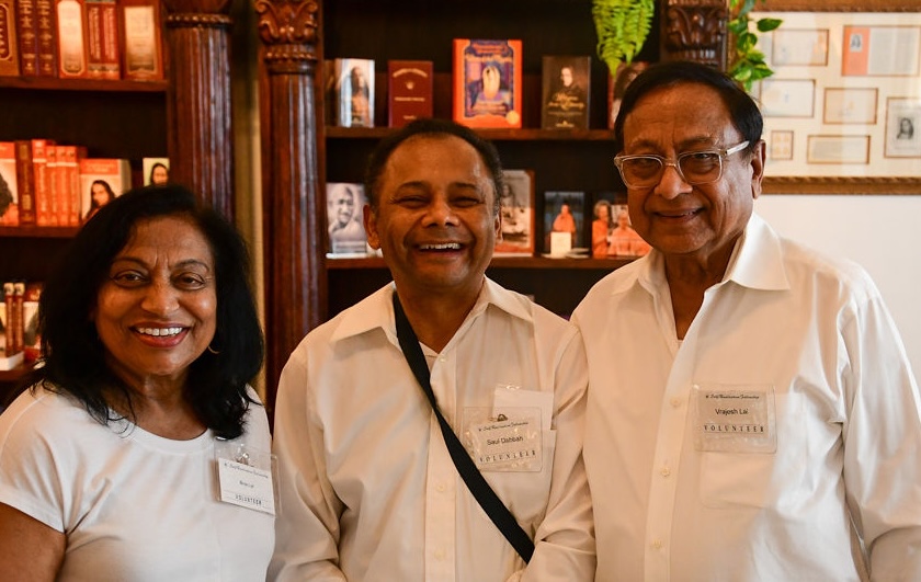 Three smiling adults wearing white shirts and volunteer badges in a room with wooden shelves and framed pictures.