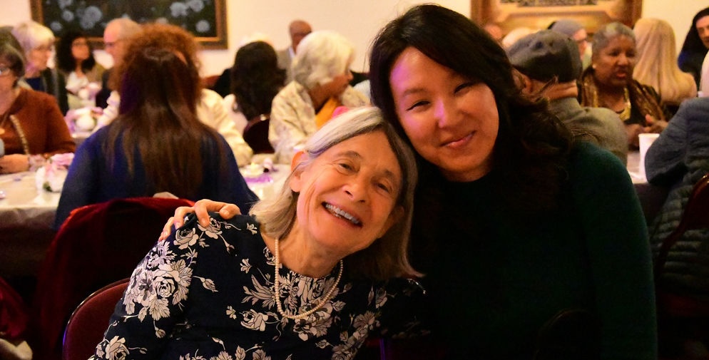 Two women smiling and hugging at an indoor social gathering with other seated people in the background.