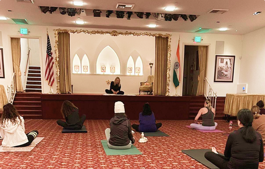 Women seated on yoga mats following instructor on stage in SRF Hollywood Temple with flags and framed photos.