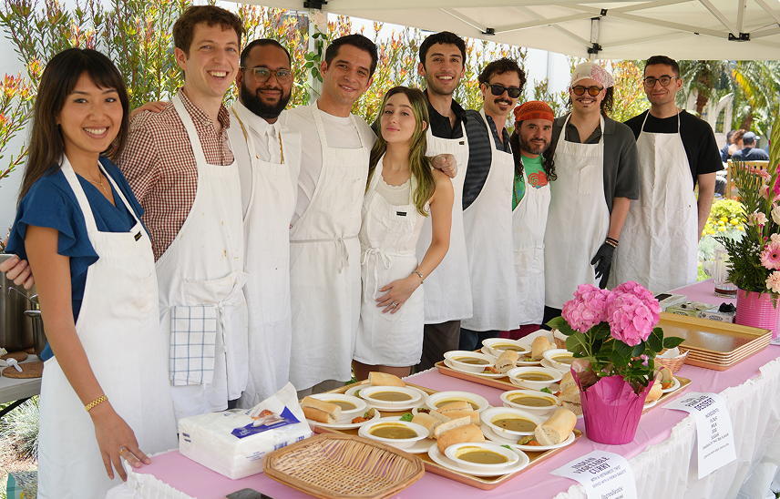 Group of smiling volunteers wearing white aprons standing behind table with soup bowls and bread under canopy.