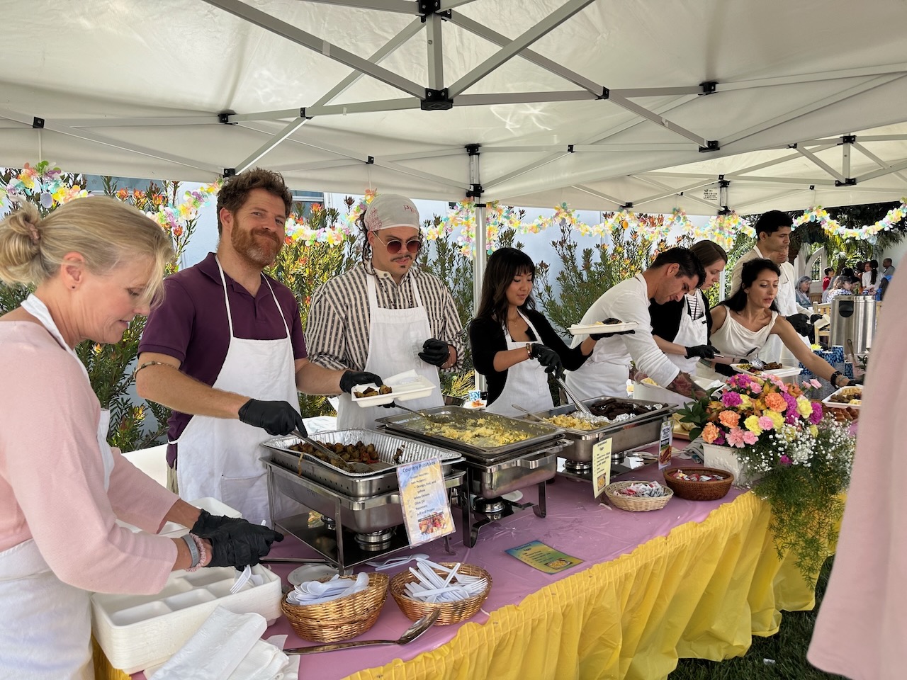 Volunteers serving food from chafing dishes under a white canopy at an outdoor event table with flowers.