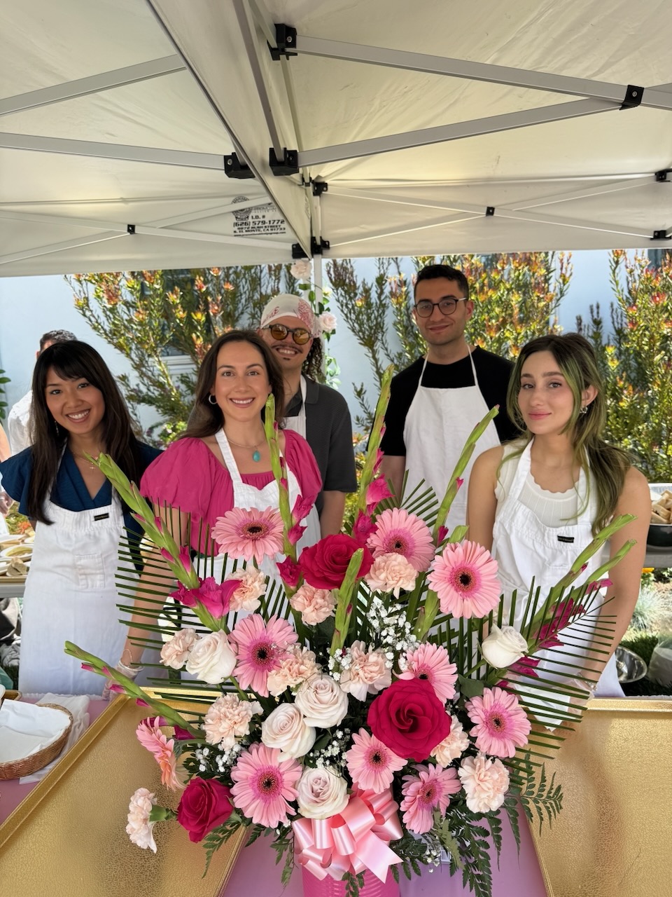 Five people in aprons behind a large pink and red floral arrangement under a canopy tent outdoors.