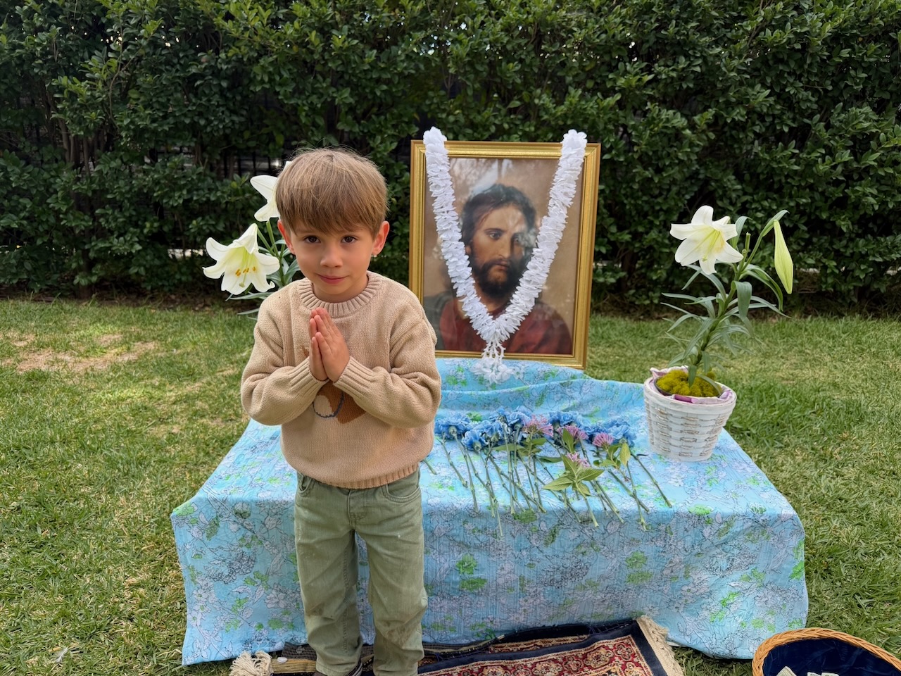 Young boy with hands clasped in prayer standing outside near a table with flowers and a garlanded portrait.