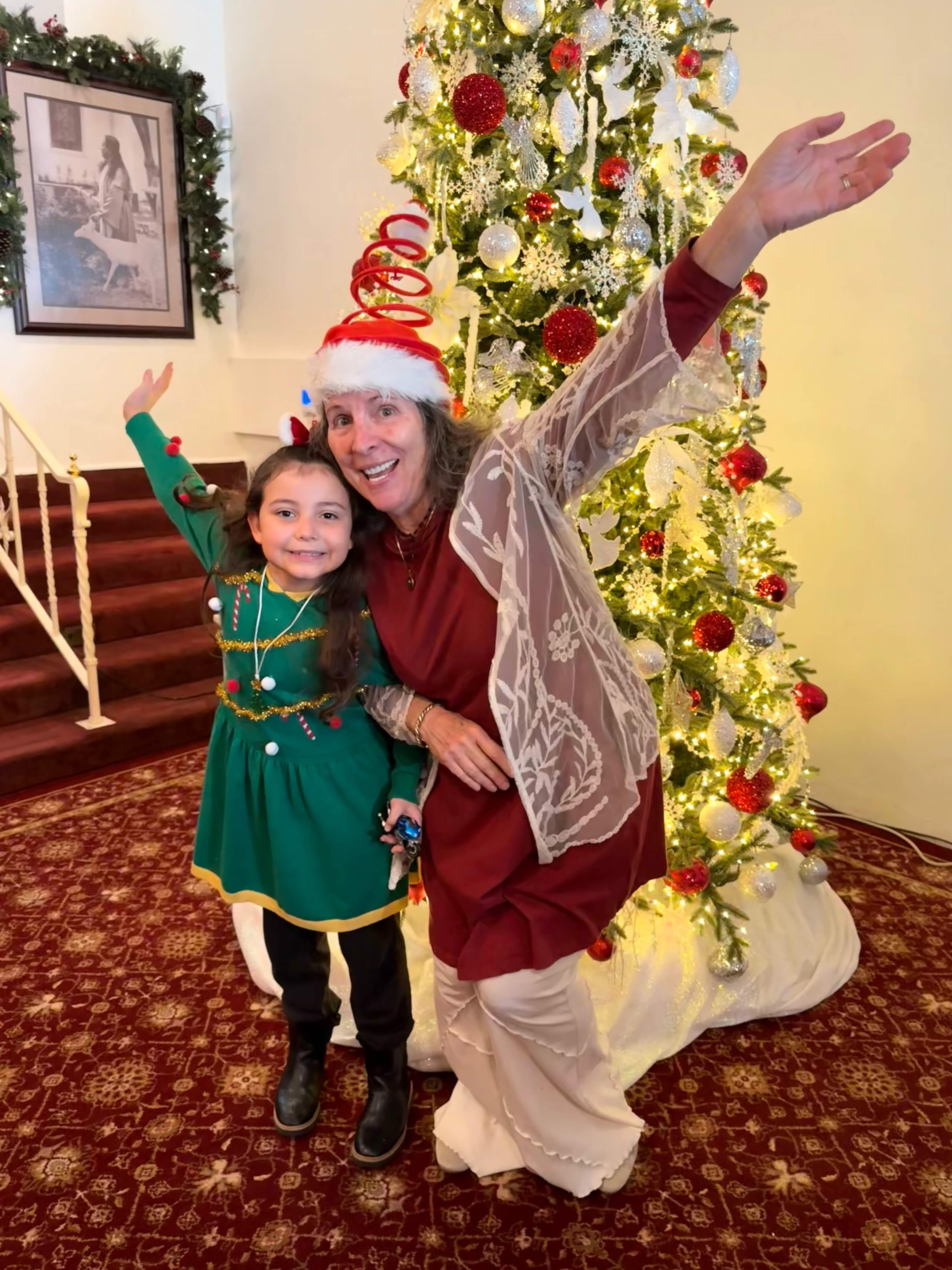 Smiling woman in Santa hat and girl in green holiday dress posing cheerfully by decorated Christmas tree.