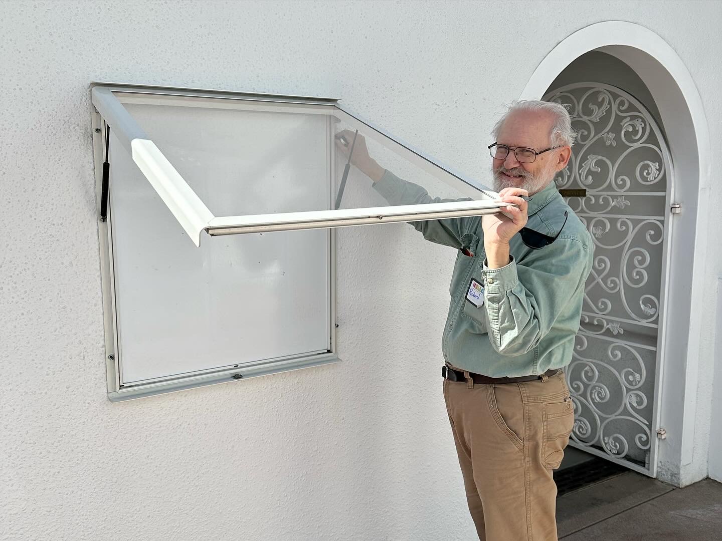 Smiling elderly man opening a clear outdoor display case mounted on a white wall near a decorative gate.