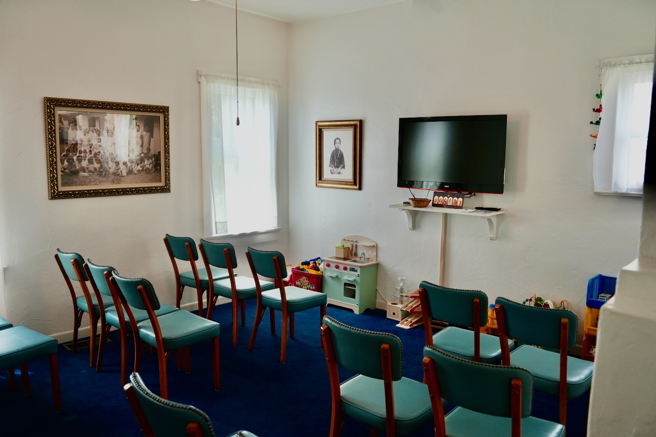 Room with teal chairs arranged in rows facing a TV, framed photos, and children's toys against white walls.