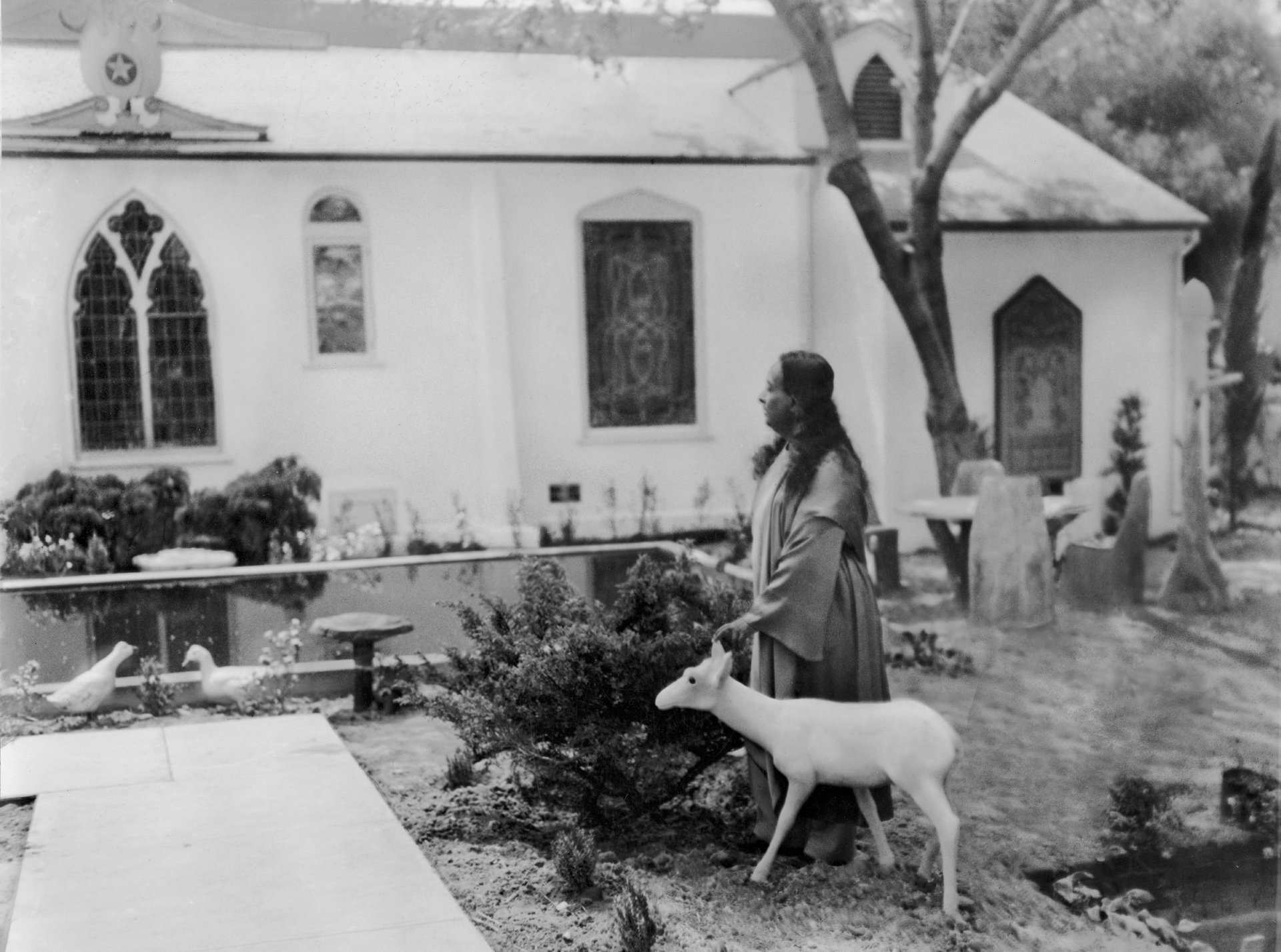 Paramahansa Yoganand standing in a garden holding a white deer sculpture near a building with stained glass windows.