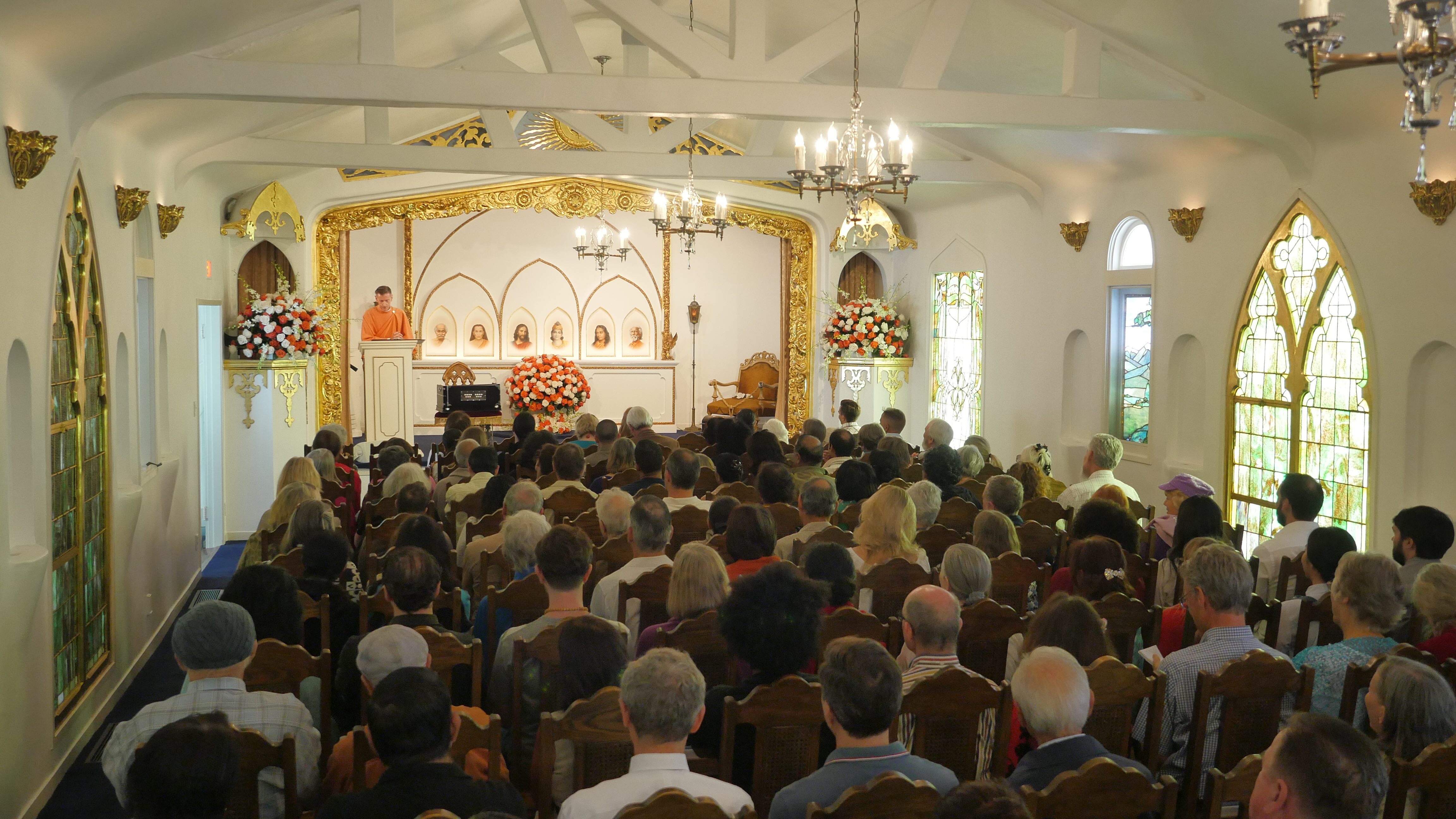 Large congregation gathered in ornate white temple with golden altar and chandeliers