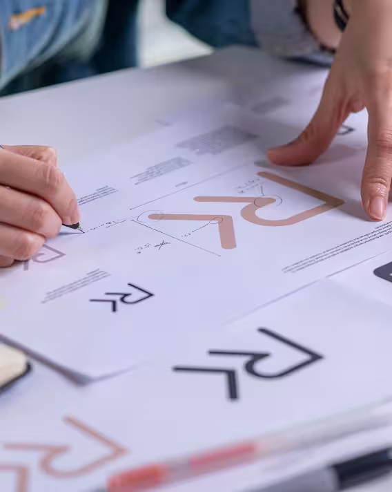 A designer sketching logo concepts on paper beside a laptop and color swatches.