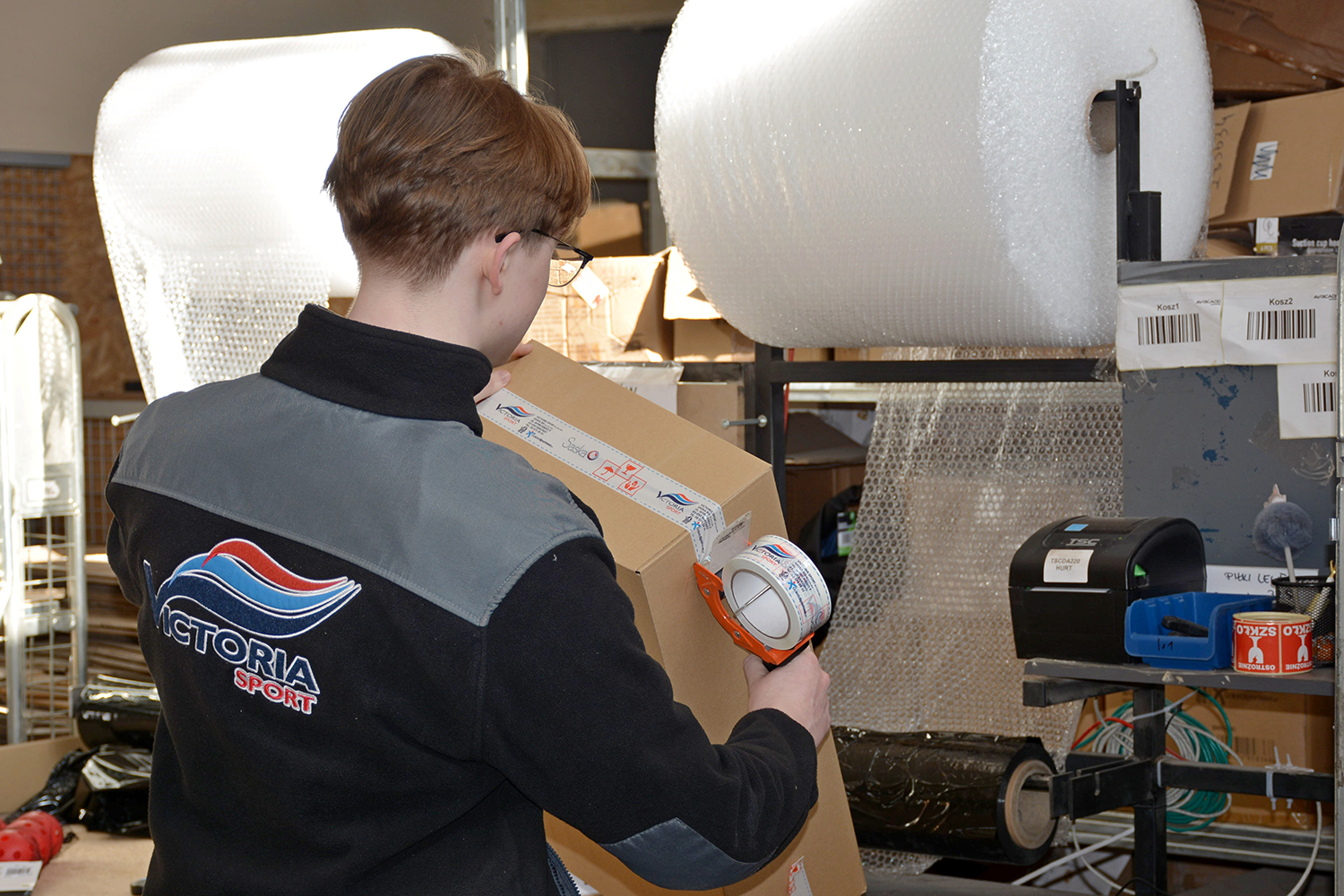 Pracownik w kurtce z logo Victoria Sport pakuje kartonowe pudełko taśmą pakową w magazynie z materiałami do pakowania.