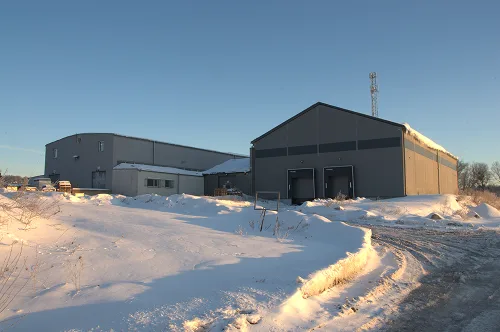 Dwa duże magazyny przemysłowe otoczone śniegiem pod jasnym, błękitnym niebem podczas zachodu słońca.