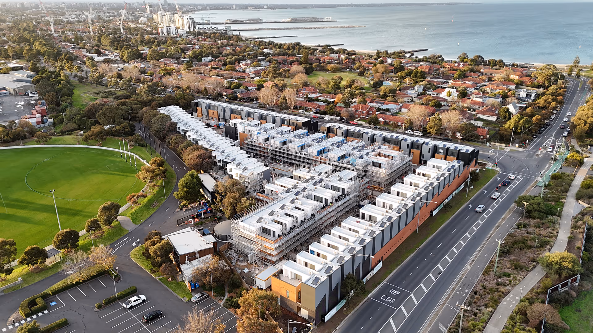 Aerial view of buildings and roads with the ocean on one end of the image and a football ground on the other