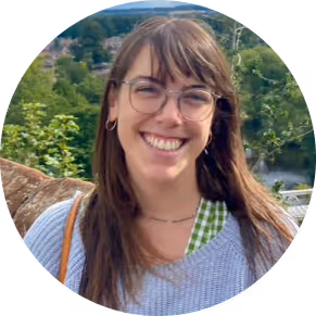 Smiling woman with long brown hair, glasses, and hoop earrings standing outdoors with trees and houses in the background.