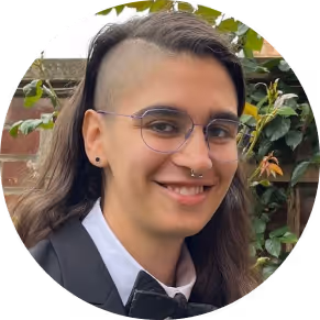 Person with long dark hair, glasses, and a nose ring smiling outdoors with greenery and a wooden fence in the background.