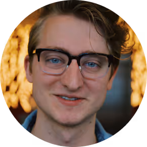 Young man with glasses and blue eyes smiling, with blurred warm lights in the background.