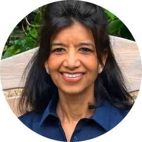 Smiling woman with long dark hair wearing a navy blue shirt sitting outdoors in front of greenery.