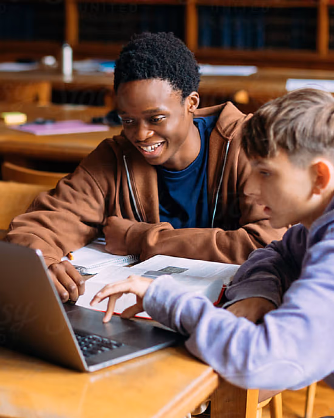 Two students sitting at a wooden table, looking at a laptop and smiling while studying together with open books.