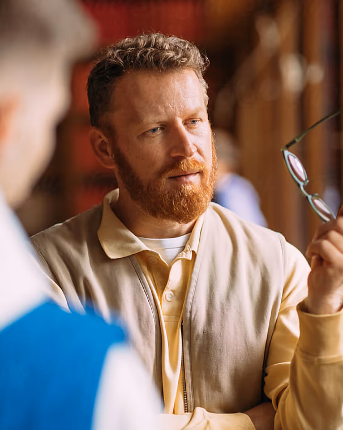 Man with curly hair and red beard holding eyeglasses, engaged in conversation indoors.