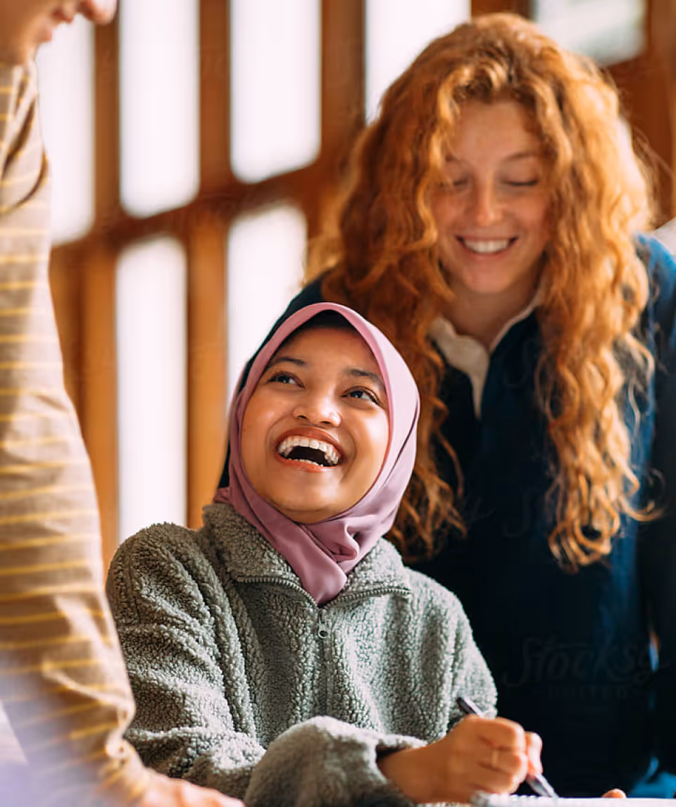 Smiling woman wearing a hijab and fleece jacket interacting with two other people in a warm, sunlit room.