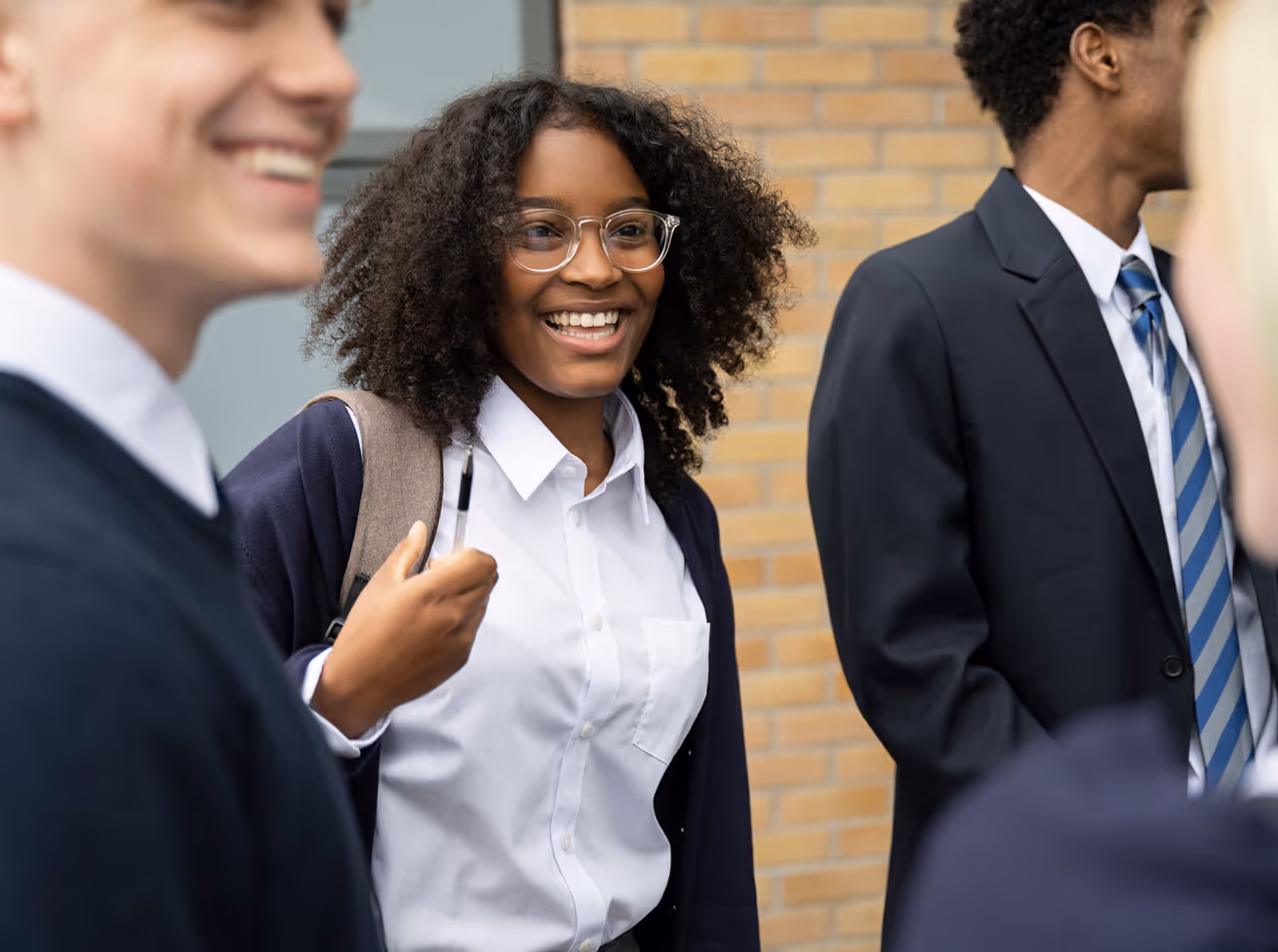 Smiling female student with curly hair and glasses talking with peers outside a brick building.
