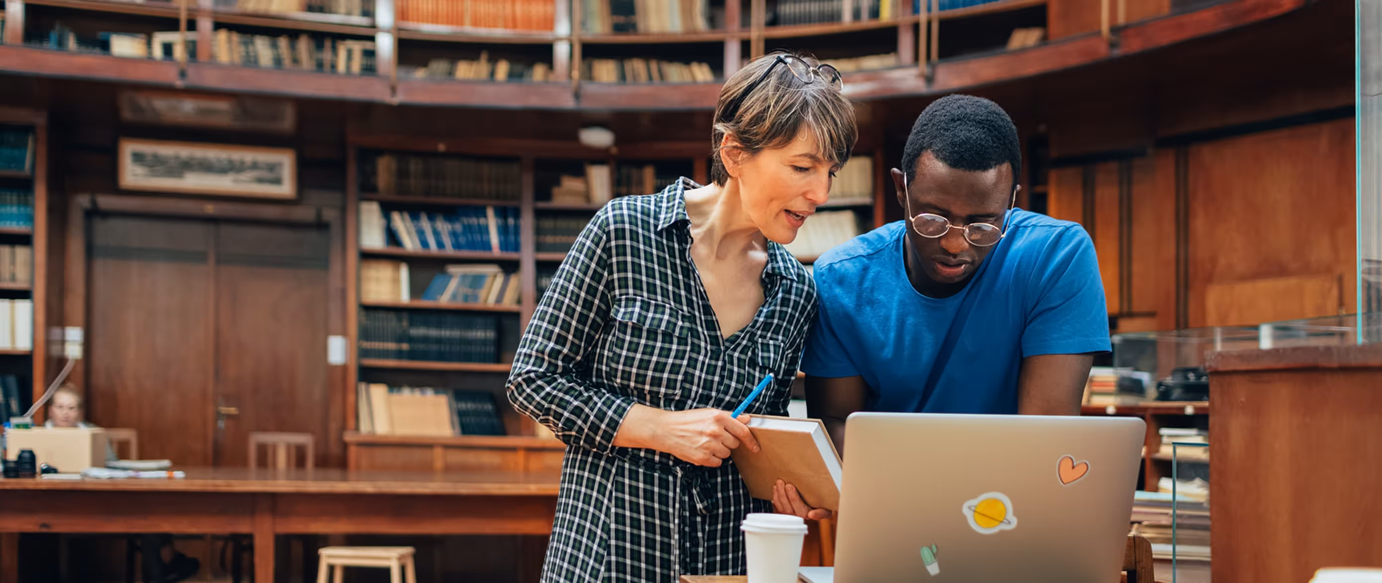 Two people collaborate in a library, looking at a laptop with stickers while holding a book and pen.