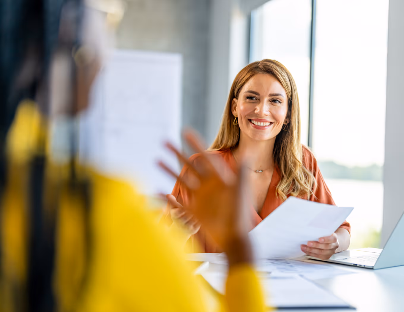 Smiling woman holding papers and talking to another person in a bright office.
