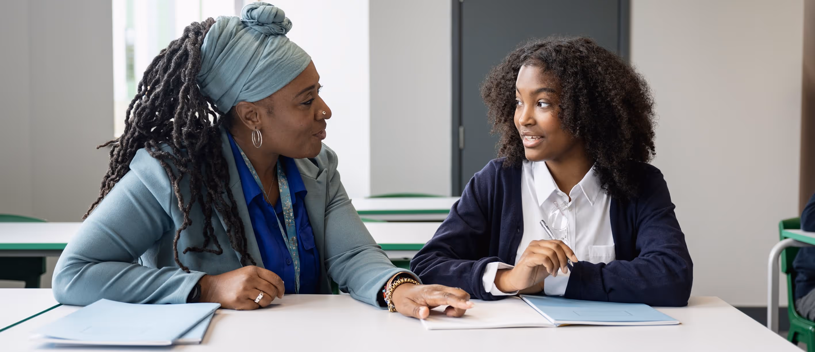 Teacher and student sitting at a desk in a classroom, engaged in a conversation with notebooks open in front of them.