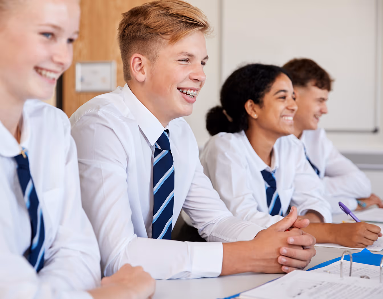 Four smiling high school students in uniforms sitting at desks in a classroom.