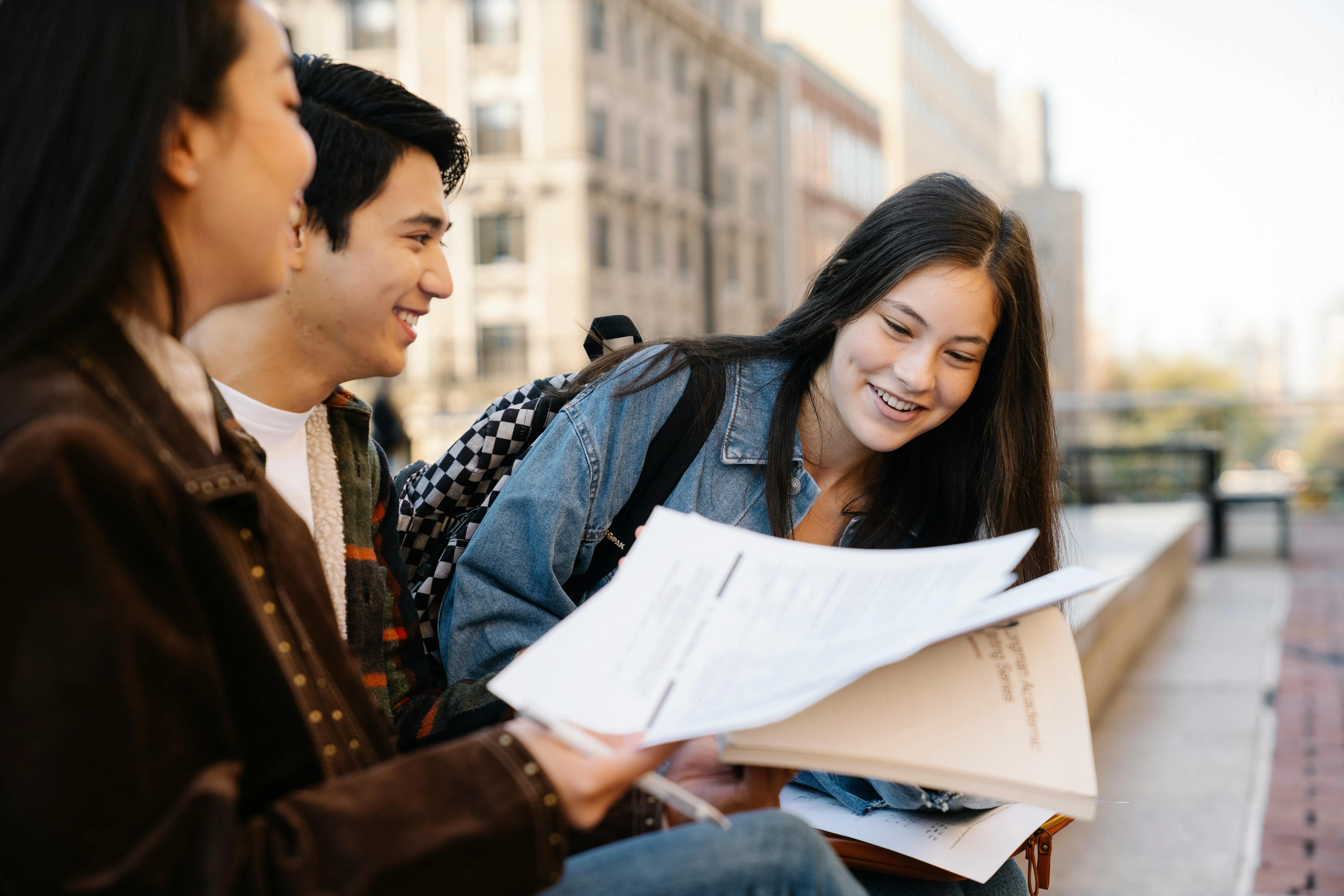 Three high school students smiling as they review college application papers outdoors on a school campus.
