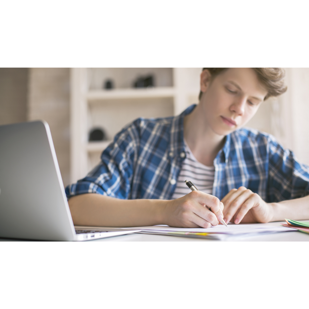A teenage boy sits at a desk near a laptop, writing in a notebook.