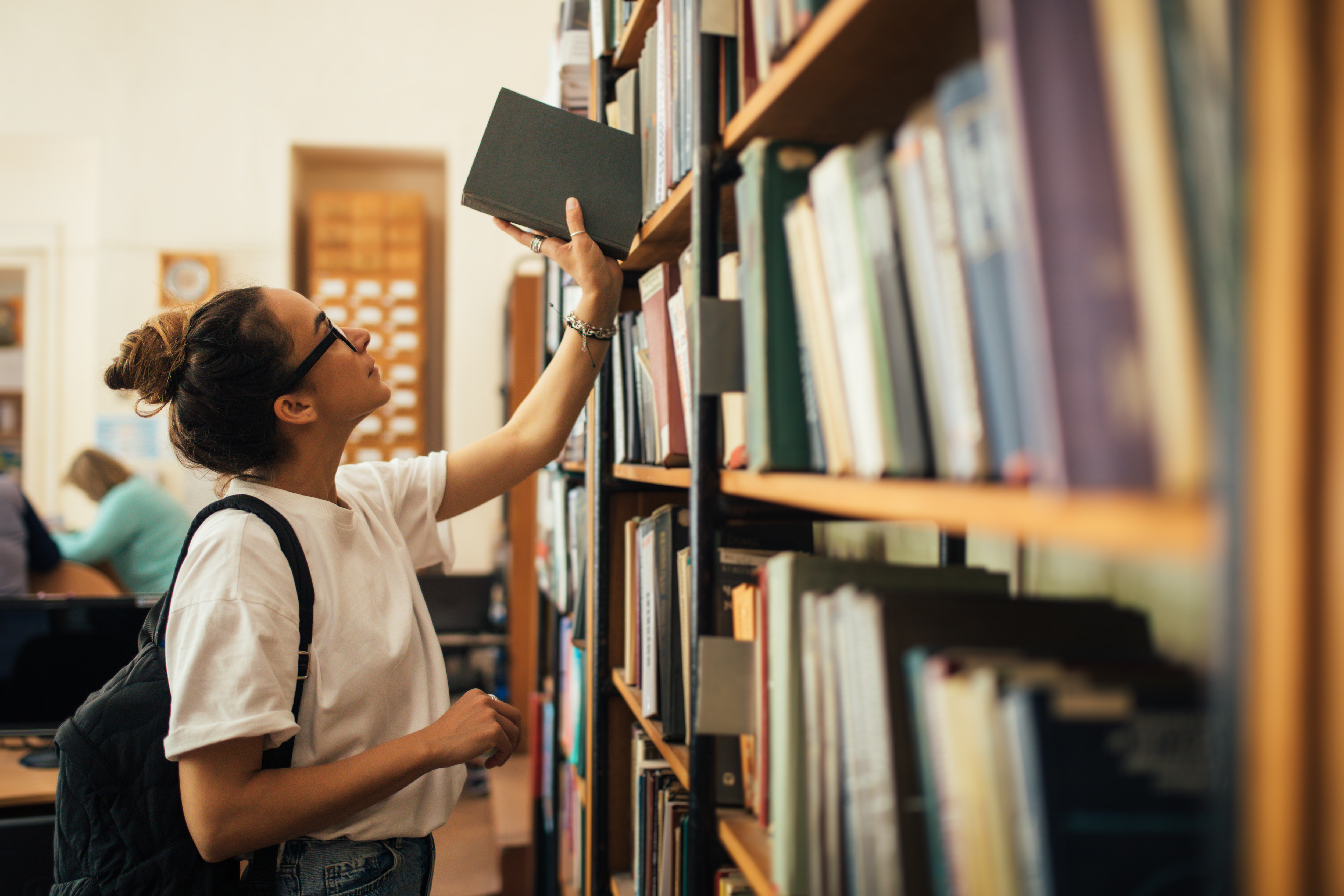 A teenage girl takes a book off a library shelf. 