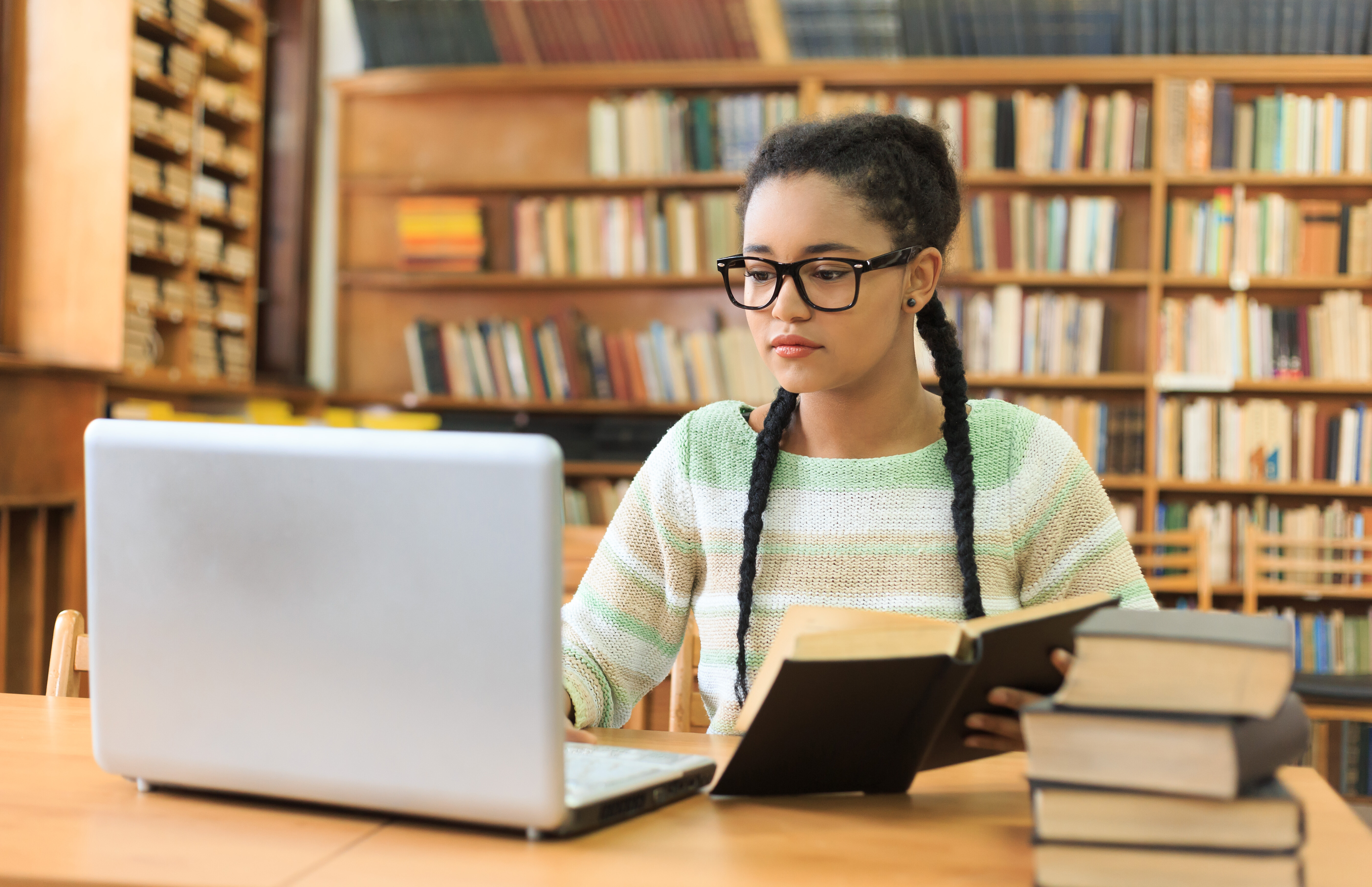 A teenage girl with plaits reads and looks at a laptop in a library. 