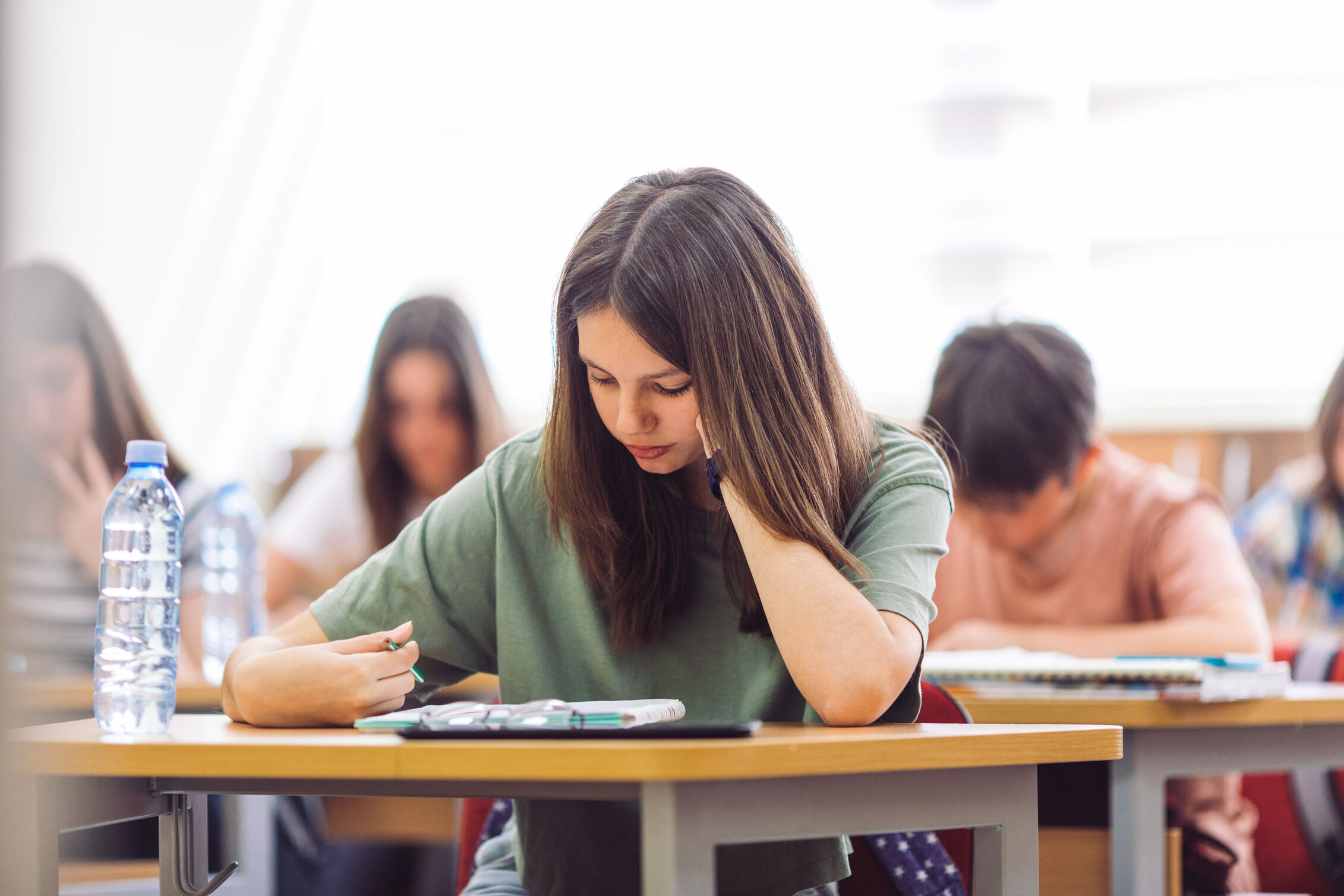 A brunette teenage girl, two classmates in the background, takes a test at a desk.