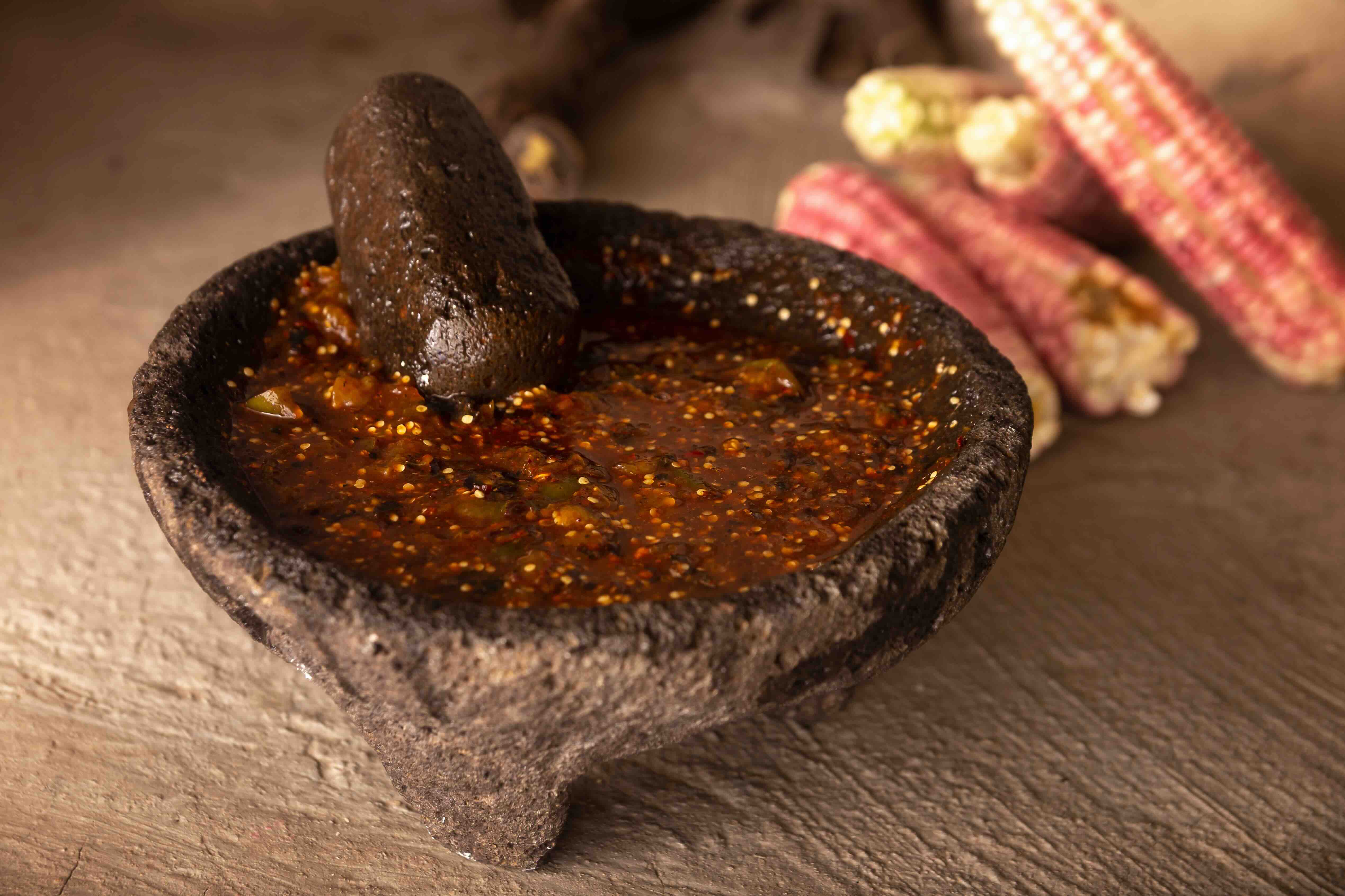 Fresh red salsa sitting in a volcanic rock molcajete. A red variety of dried corn rests in the background.