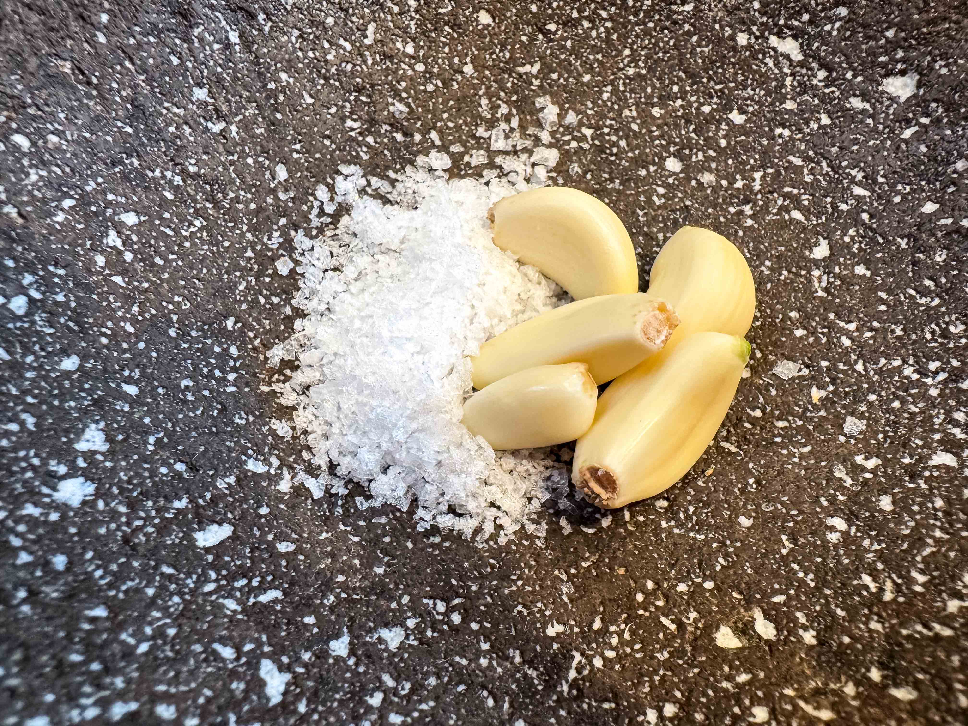 A close-up of the molcajete. Some salt and a few cloves of garlic rest inside. Tiny bits of white rice powder are compacted into the porous surface of the molcajete.