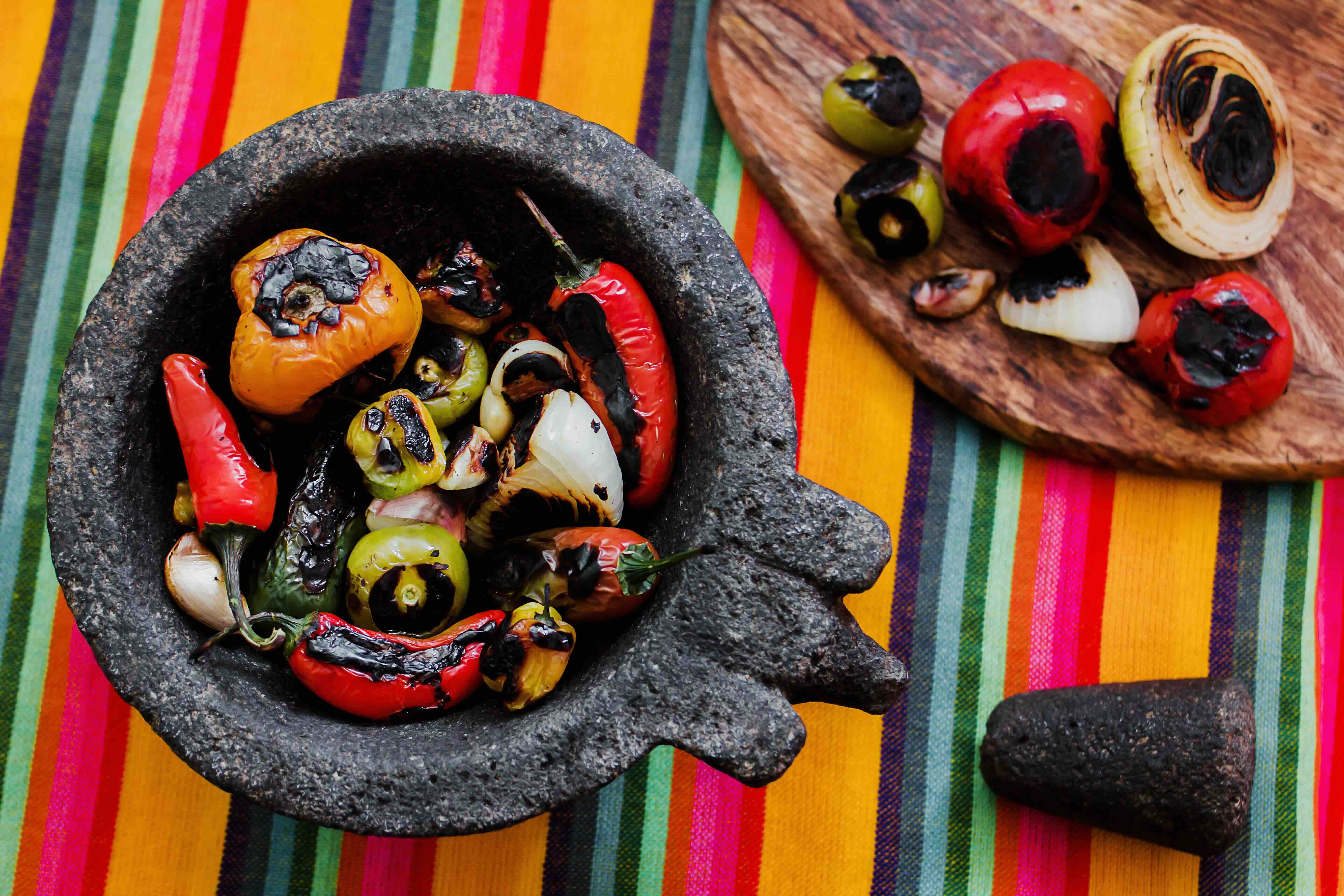 Roasted tomatoes, tomatillos, onion, garlic, and bell pepper inside of a molcajete, sitting on top of a colorful striped tablecloth.