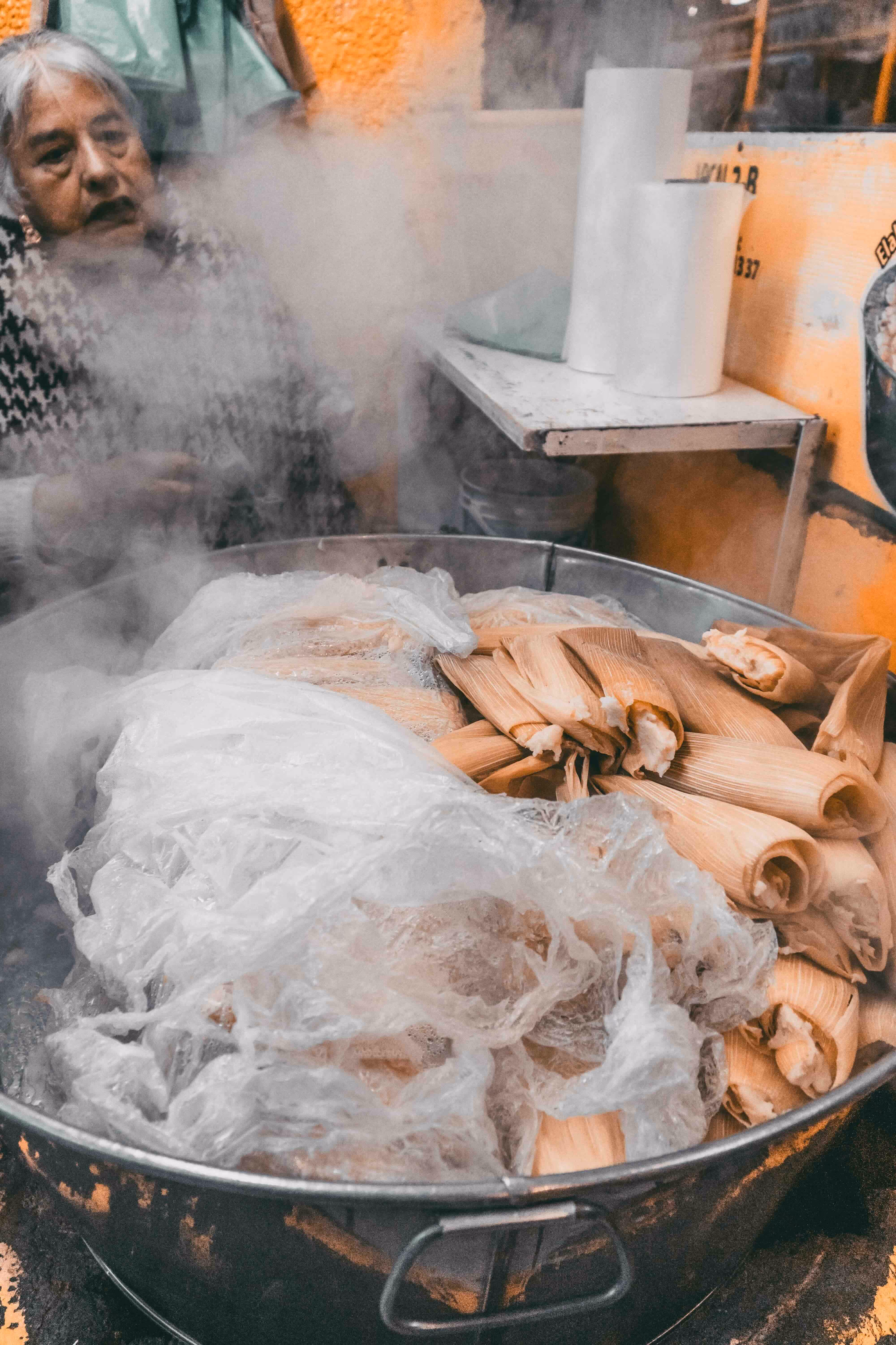 A woman standing behind a huge steel pot of steaming tamales.