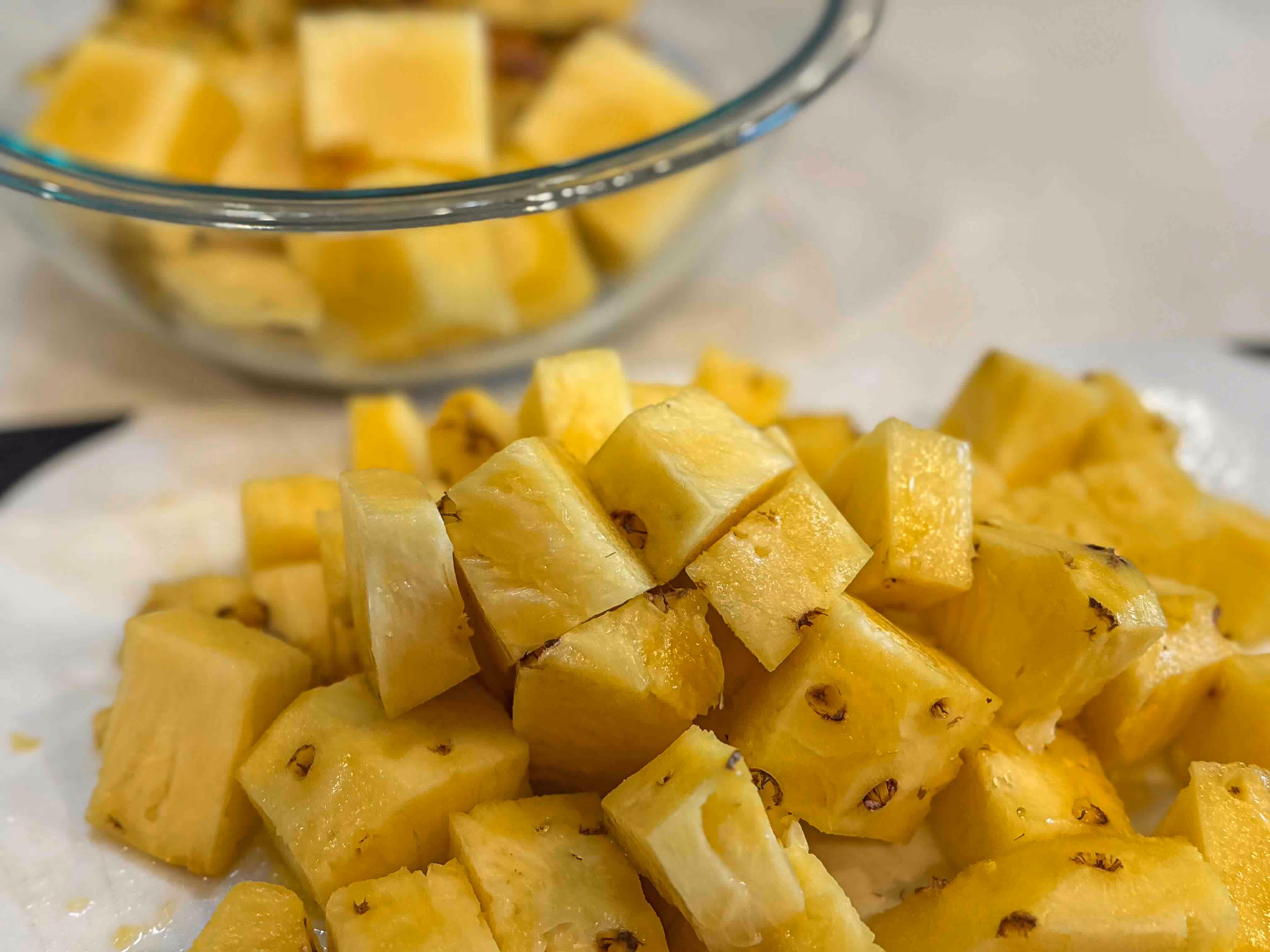 Cubes of fresh-cut pineapple on a cutting board. The core and peel are in a bowl in the background.